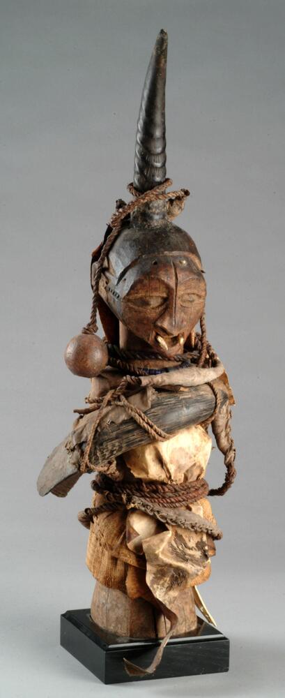 This imposing, anthropomorphic Songye <em>nkisi</em>, or power figure, stands upright and features an assemblage of man-made, vegetal, and animal components. Most strikingly, a large antelope horn protrudes vertically from the figure’s head, and two animal teeth have been affixed to the corners of its mouth giving the appearance of fangs. Bearing wide, alert eyes, this fearsome figure is enswathed by civet and monkey skin. Horns, including a smaller one inserted into a larger one, have been tightly strapped around the figure’s chest. Also attached to the figure are seed pods, an ax blade, and glass beads. A medicine bundle carrying feathers and a horn hangs behind the figure's back.
