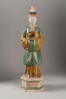 This earthenware sancai-glazed figure of a male attendant is standing on an octagonal cream and green dais, wearing a long green tunic over an amber-colored underrobe, with amber and green sleeves, carrying an amber-glazed bowl. His hair, hat and face are painted with polychrome mineral pigments, and his tall hat painted to match his garments in an orange-amber color. The head has been sculpted and painted separately from the body.
