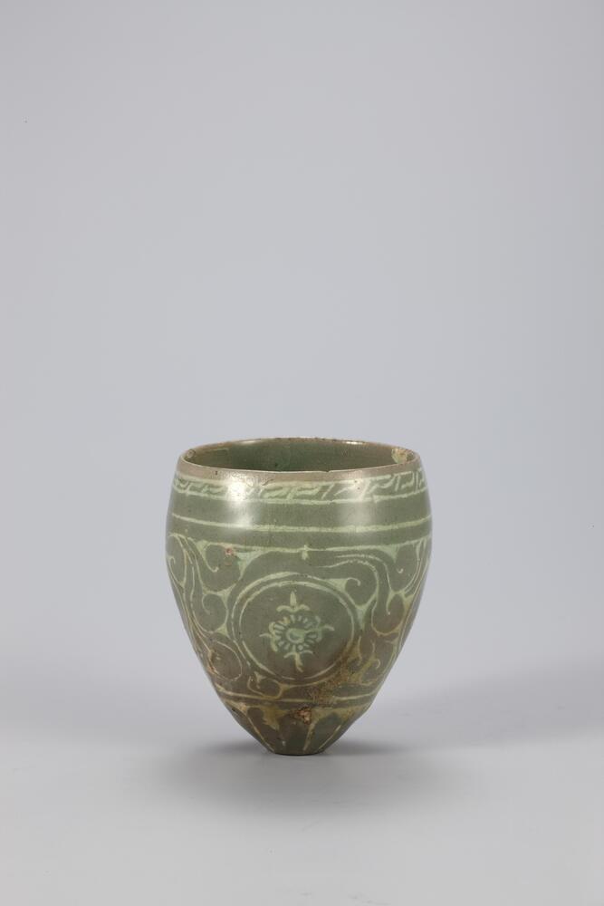 Earthenware stirrup cup with sgraffito design and celadon glaze. A chrysanthemum blossom decorates the tapered base of the cup, and widens into a large middle register with four stylized chrysanthemum medallions. The cup curves back inward toward the rim of the cup, decorated with a fret pattern just below.
<p>This is a conical stirrup cup designed to be inserted into a cup holder. It is a fine piece of celadon with the sudtle beauty of white inlaid decorations in match with the blue-gray body. Its entire outer wall is decorated with inlaid motifs of fret, chrysanthemums, scrolls and lotus petals.<br />
[<em>Korean Collection, University of Michigan Museum of Art</em> (2014) p. 128]</p>
