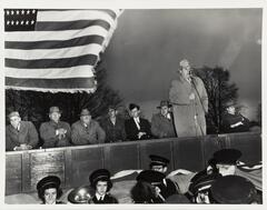 A black and white image of a man speaking into a microphone with seven men seated behind him. A band is in the lower center foreground and an American flag is in the upper foreground. 