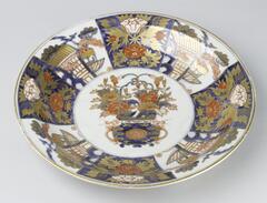 A deep plate featuring a design of red and gold flowers, with a blue pot in the center, and smaller designs of buildings and flowers against backgrounds of white and blue.  On the underside of the plate, in the center, is a small red flower.  