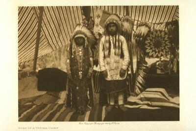 This is a portrait of two young boys. They stand inside a tipi or tent, wearing large headdresses and beadwork around their shoulders. The interior of the space is filled with patterned blankets and cloths. 