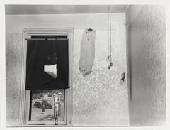 A black and white image of the interior of a room with a window facing a street or parking lot. The wallpaper is peeling and stained and the windowshade is torn. 