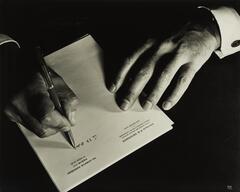 A black-and-white photograph of two hands, one holding a pen and writing on notepaper, the other placed on the paper to the right. The letterhead on notepaper is for the office of &quot;Doctor P.E. Seidmann&quot;.