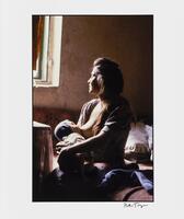 A color photograph of a woman breastfeeding. She is seated near a window and looks out toward it.