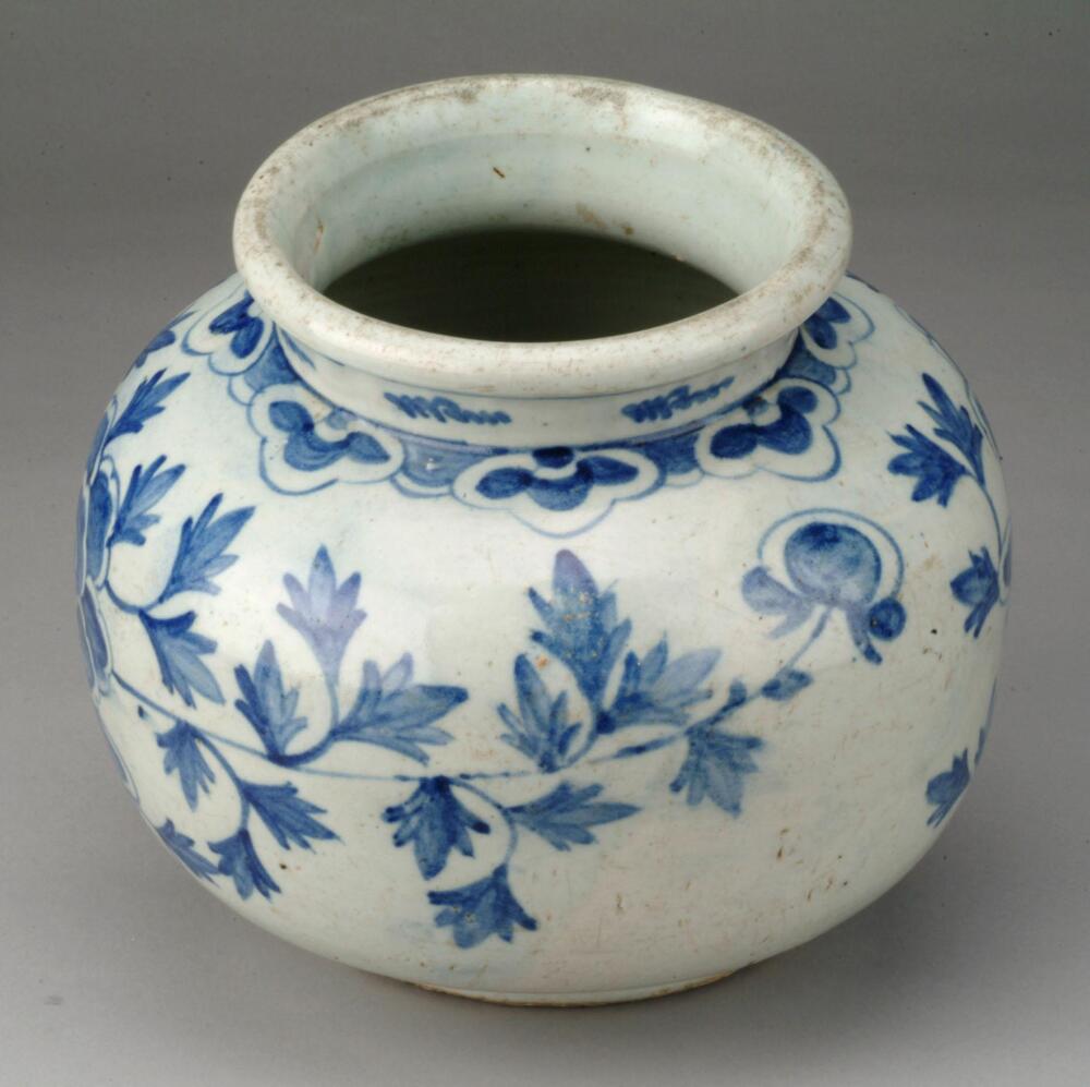 This jar is assumed to have been produced near the Bunwon-ri kiln, Joseon&rsquo;s last official court kiln, after its privatization. Its shape and design follow the blue-and-white porcelain produced in Bunwon-ri kiln, although the jar was warped during firing. It features a foliage design around the neck, a yeoui-head design around its shoulder, and peony scrolls on the belly, all painted in cobalt blue. The jar has short neck and flared rim. The jar is glazed all the way down to the foot rim, on which remain sand spur marks. Its background color is dark, while the pigment has spread in some parts.<br />
[Korean Collection, University of Michigan Museum of Art (2014) p.172]