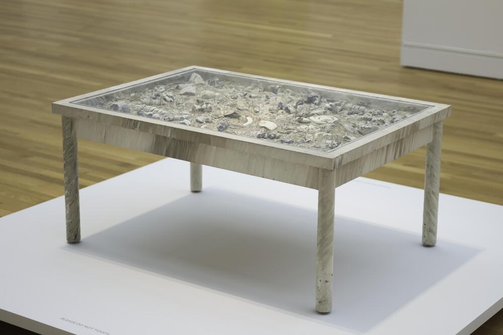 A table with painted metal legs and frame. The top is covered in shells, some gold-painted. A glass top covers the surface of the table.
