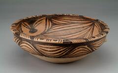 A wide round bowl with an articulated, everted wide rim and a conical lower body on a flat base. It is decorated with wavy linear patterns on the upper half of the exterior, and swirling lines with dots and networks of cross-hatching on the interior. Cross-hatching, curved lines and dots are on the rim.  