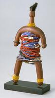 Carved wooden human figure with a columnar body and limbs. The body is decorated with strands of beads, primarily red, white, and purple. The neck and ankles have multiple strands of yellow beads while one wrist has a metal bracelet. The top of the figure is decorated with human hair.&nbsp;