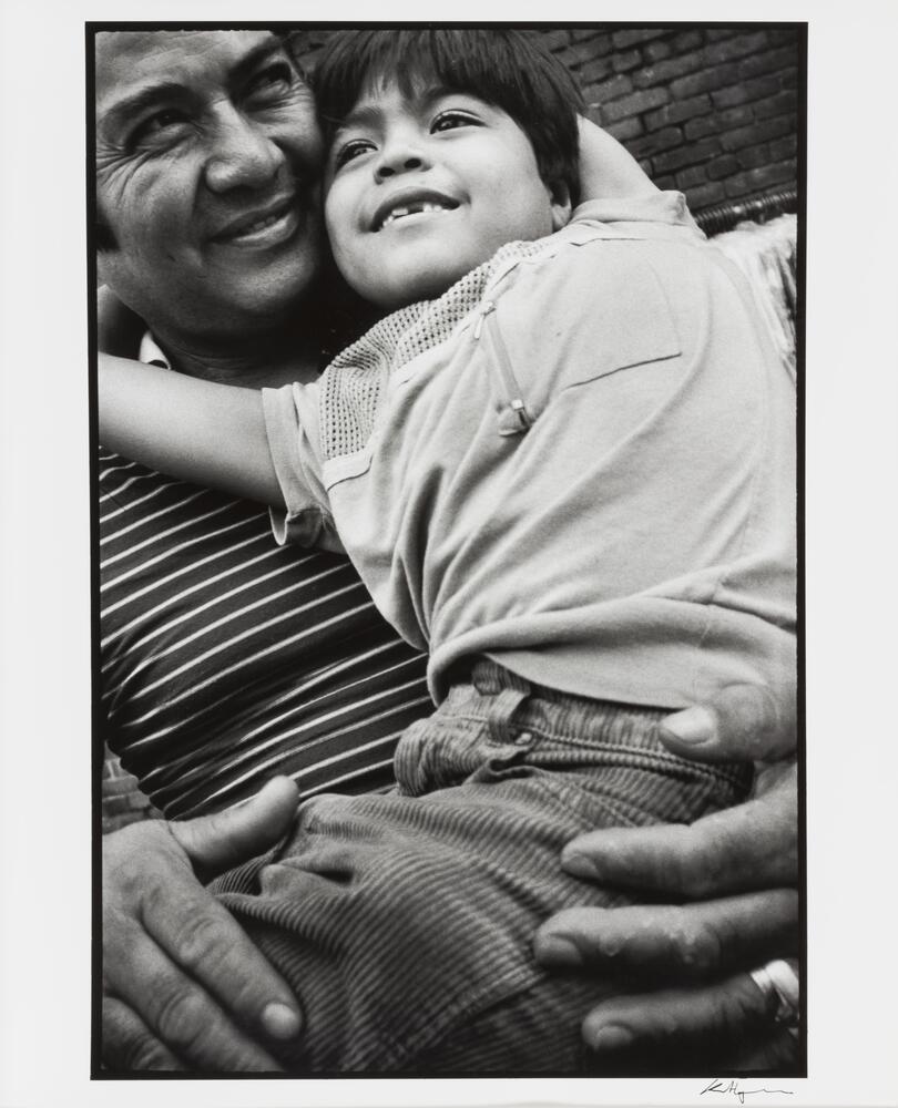 A black and white photograph of a man holding a young boy in his lap, the boy looking forward. The boy&#39;s arms reach up and around the man&#39;s neck, and the man looks out and to his left.