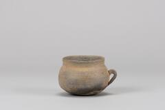 It has a flat base, globular body and straight neck. There is no design on the surface of the body. The attached handle is a little small and thick.<br />
<br />
This is a gray, high-fired stoneware cup with a handle. Its mouth is completely upright, and its rim has a sharp edge. The section immdiately below the mouth tapers inwards and is then connected to the round body. The body is widest at the center. The handle is attached to the lower-central part of the body; the upper end of the handle penetrates the side of the cup, while the lower end is joined by simply rubbing it against the cup&rsquo;s surface. There are traces of rotation and water smoothing on the inner and outer surfaces of the mouth. Natural glaze is visible in the parts around the round base.
<p>[Korean Collection, University of Michigan Museum of Art (2017) p. 68]</p>
<br />
&nbsp;