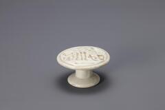 A small glazed porcelain piece resembling a cake stand with a narrow stand and wide-lipped base. The top is detailed with a carved pattern that would imprint the rice cake being molded on it.<br />
<br />
Rice cake stamps are used to impress designs upon rice cakes. They are generally made of wood or ceramic. Ceramic rice cake stamps normally come in the form of round stamps and consist of a patterned surface and a handle. Patterns, carved or raised, on the stamp vary from geometric lines to auspicious designs that wish for prosperity and longevity. Their small size makes them highly portable, while their simple yet contemporary designs have mad them popular among collectors. The University of Michigan Museum of Art collection includes nine white porcelain rice cake stamps. Some are gifts from Mr. and Mrs. Hasenkamp, and others are gifts from Ok Ja Chang and the Chang family.<br />
<br />
The Chinese characters &ldquo;壽&rdquo; (&ldquo;su;&rdquo; longevity)&rdquo; and &ldquo;福&rdquo; (&ldquo;bok;&rdquo; ha