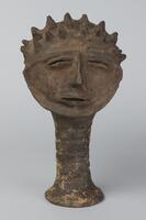 Ceramic head on a hollow cylinder with a fluted base. The figure&#39;s eyes and mouth are thin, while the nose is angular. The top of the head is covered in small protruding points.&nbsp;