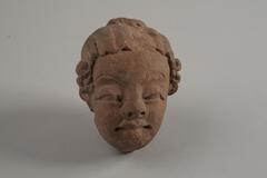 An Indian terracotta of a head figure of a young child, which has distinguishable facial features and facial expressions. The head figure is biting the lips which seems to shows fear and nervousness.