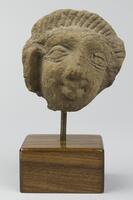 A stone head with joined eyebrows, vertical incisions to represent stylized hair, a large lobed right ear, and a smile.