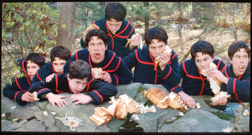A photograph of eight boys, all of which are the same person, voraciously eating a loaf of white bread off a rock in a forest.