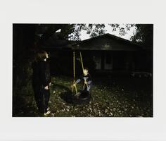 An image of two children in a front yard, both wearing dark clothing. One child holds the yellow chain of a tire swing, while the other looks upward. A dark tree and house stand in the background.