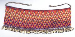 An apron made from red, blue, white, and yellow beads in a zig-zag pattern. There are two strings attached at the top of the apron, to tie around the waist. The bottom of the apron is decorated with a fringe of cowrie shells.&nbsp;