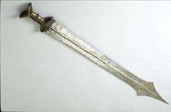 This sword has a long blade with a tri-pointed tip. The center of the blade has a ridge running the length of it. The wooden handle has a V-shaped top.