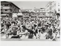 A crowd of people, many wearing bathing suits, sit viewing a platform. Behind them are storefronts and a Coca-Cola sign.&nbsp;
