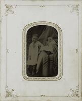 A sepia-toned image of two children and a larger, seated figure draped in a blanket. The smaller child is an infant that sits in the lap of the seated figure and the larger child stands to the infant's right with an arm on the seated figure's leg. 