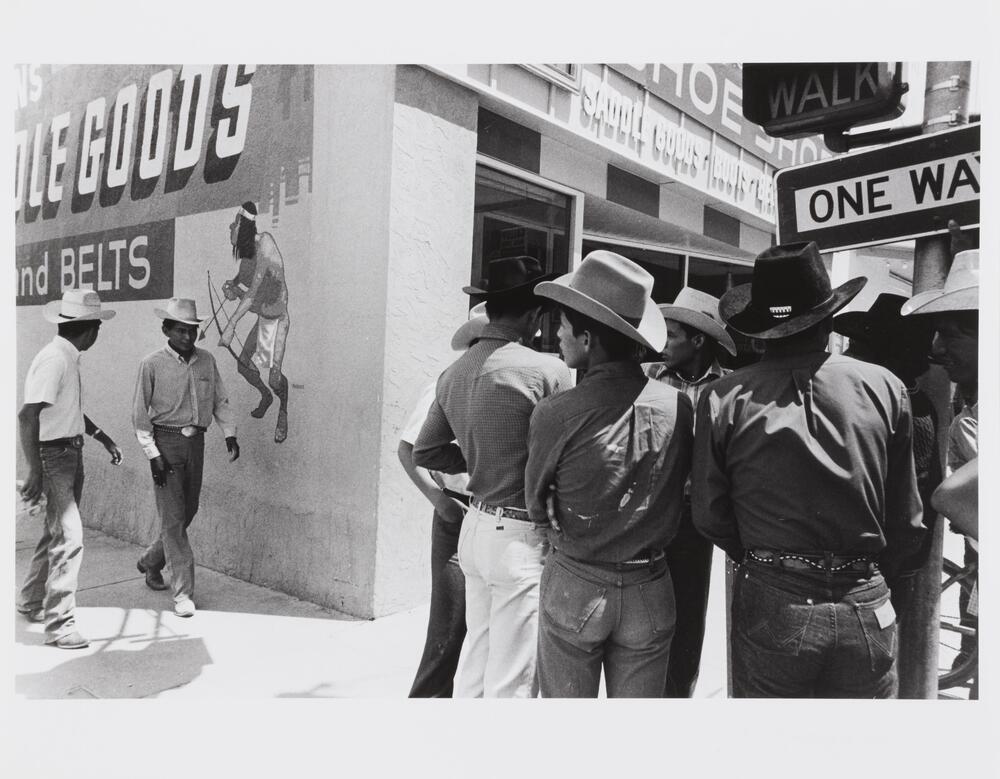 Photograph of a group of men wearing jeans and cowboy boots and hats congregating on a street corner. 
