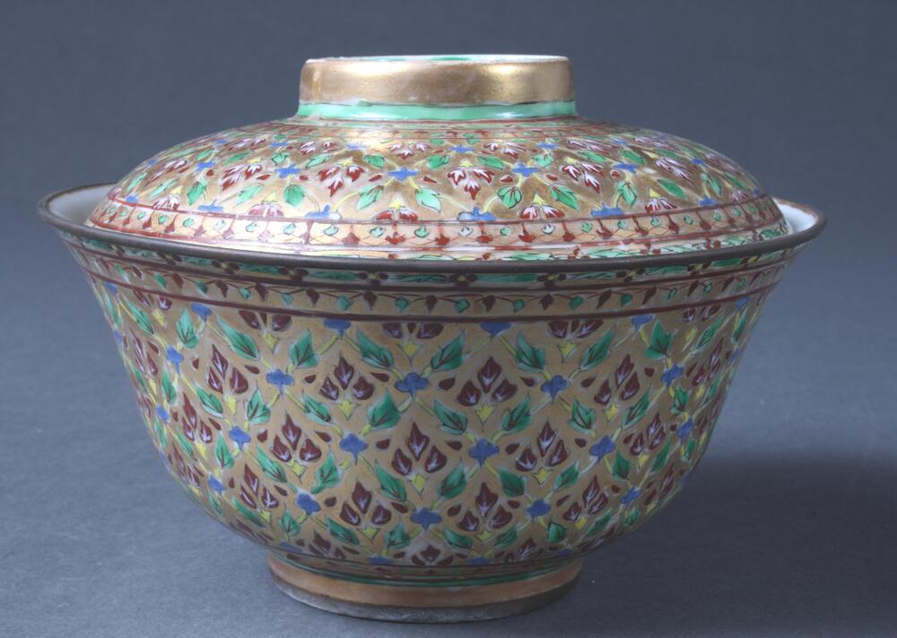 Covered ceramic jar with brilliantly colored overglaze enamel painting in repreating floral patterns.  A small base curves into a deep, wide bowl with a flaring mouth, in which the lid rests.  The lid can be turned upside down to provide a shallow bowl, the base of which (or topmost portion of the lid) is encircled by gold and green bands.