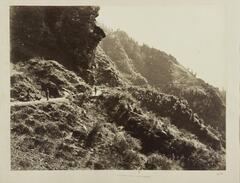 A black and white image of a road carved into steep, rocky hills. &nbsp;The hills are covered in shrub-like vegetation. &nbsp;Four figures are visible on the road. &nbsp;Three are positioned near the viewer, and the fourth walks further up the road, away from the viewer.