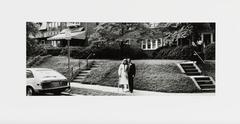 A black-and-white photograph of a couple standing on a sidewalk in front of a row of brick houses. A car, to the left, has "Just Married" written on its rear window.