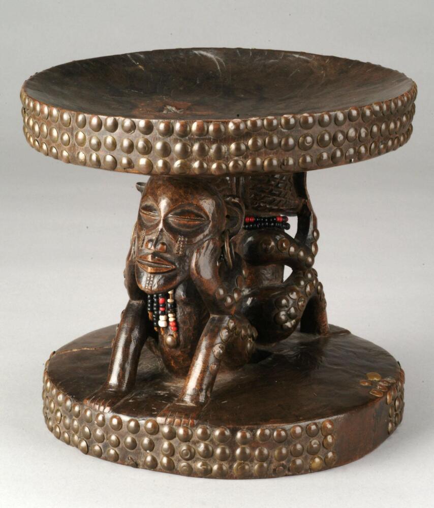 This hour-glass shaped stool is supported by two caryatid figures who sit in a pose of lamentation&mdash;crouched with head in hands. Scarified patterned abstracted tears spill from their lower eyelids. Brass studs adorn the perimeter of the stool&rsquo;s seat, base, and figures. Both figures wear strings of black, red and white beads around their necks.