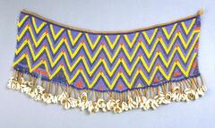 An apron made from multi-colored beadwork, primarily red, blue, and yellow beads in a zig-zag pattern. The bottom of the apron has a fringe of cowrie shells. 