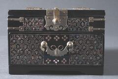 This mirror box is decorated with lacquer and mother-of-pearl inlaid designs. Thelid of the box opens upwards, and has brass hinges and a closure in the sahpe of a flower. The lower drawer has a handle in the shape of a bat.<br />
<br />
This box has a mirror attached to the inside of its lid and was used by women. The box is decorated with hexagon and bamboo motifs by employing <em>kkeuneumjil</em> and <em>jureumjil</em> techniques. Hinges made of nickel are decorated with chiseled peonies on a ringmat ground. The corner brackets, or side wrappers, are in &ldquo;I&rdquo; shape. The single drawer has a handle in the shape of a bat, realistic to the extent of depicting antenna. Lacquer was heavily repainted on the base of the drawer. The frame of the mirror inside was decorated with fret-patterned band on left and right sides, and saw-tooth design on upper and lower sides.
<p>[Korean Collection, University of Michigan Museum of Art (2017) p. 266]</p>
