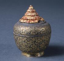 The small container with lid has a deeply engraved design filled with black enamel. The body has tight, compact, overall decoration of floral motif, with the stupa-shaped gold fitting on the top. Colored glass pieces were inlayed in the fitting.<br />