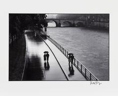 A black and white photograph of a walkway with a river to its right and bridge in the distance, Two couples walk holding umbrellas.