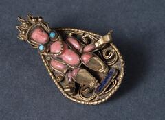 A tiny tear-drop-shaped metal (copper?) pendant, edged with beading, serving as support for a minature seated image of a Dhyani Buddha, made of inlaid semi-precious stones and copper wire.  This may have originally been part of a necklace or tiara, or part of a brooch for a high-ranking monk's ritual costume.