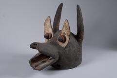 A wooden mask with zoomorphic features. The mouth is in the form of open, elongated lips. The eyes are formed by white triangular projections with small central knobs. Behind the eyes, at the back of the head, are two pointed projections that curve slightly inward. 