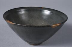 A conical bowl on a tall straight foot ring.  It is covered in a thick dark brown-black glaze, with a fine crackle covering the glazed surfaces, and three gold lacquer repairs have been applied to losses at rim. 