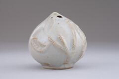 A round peach-shaped water dropper. The peach itself is covered in a white glaze and covered in bamboo stalks and leaves. These are embossed onto the peach and stand out even more as the iron brown underglaze comes through strongest on these details. The iron brown underglaze can also be seen along the base of the waterdropper. The hole is at the top of the peach.<br />
<br />
This is a peach-shaped water dropper shaped in a mold, featuring mold-impressed designs of peach leaf and branch on the surface. Its upper part is perforated by two water holes and the body is very light. Parts of the designs in high-relief are thinly glazed and tinged with brown. The foot is low. It was fired on the kiln shelf, which is an indication that it was produced in the early 20th century.<br />
[Korean Collection, University of Michigan Museum of Art (2014) p.185]