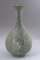 This is known as a pear shaped bottle vase with widely everted mouth, narrow neck that makes it easy to grasp and a round globular body that is bottom heavy. Five lines encircle the body and neck. The foot is rather high.<br />
<br />
Bronze bottles, bowls, plates and cutlery were placed as burial ware in Goryeo tombs along with celadon vessels. This bottle has traces of being splashed by muddy water, thus it is assumed to have been excavated from a tomb. This type of bottle with a long neck and flared mouth was also made in celadon in large quantities. The bottle is decorated with three ridges, and between the ridges are incised three thin lines. The mouth was made by folding the metal sheet inwards and joining the folds. The vertical foot has been attached separately. The entire bottle is covered by a thin patina, and part of its body has been ruptured. It, however, retains its original form and has been preserved well. Part of one side, which has been in contact with earth, is more decayed than the rest.