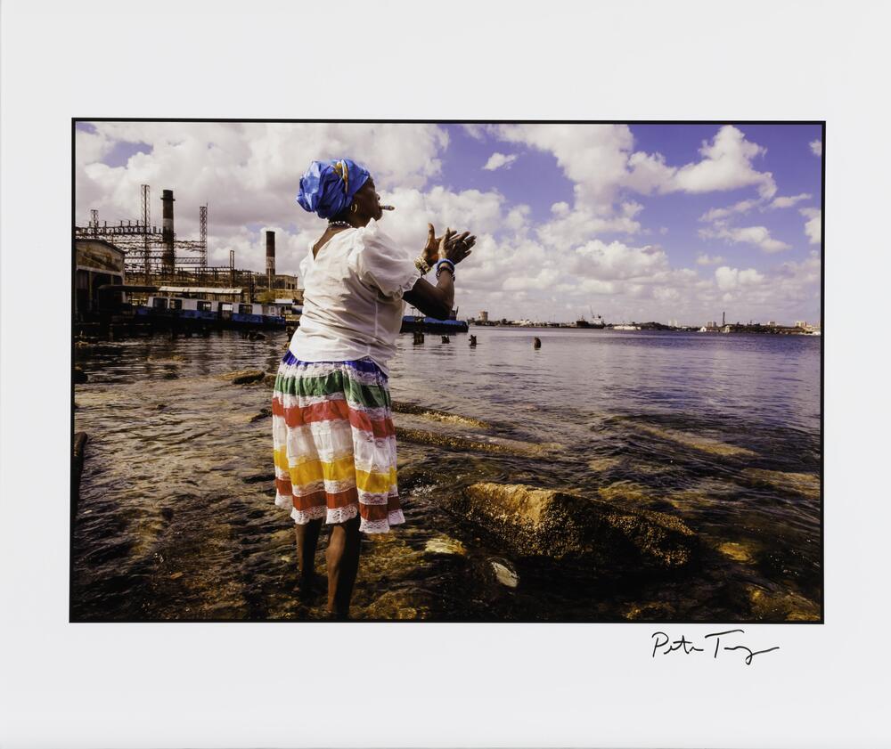 An image of a woman wearing a brightly colored head wrap and dress. She looks toward a body of water with a city visible on the opposite shore. Docks are visible to her left. She extends her hands, as if clapping.