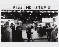 Photograph of a man and woman, surrounded by pedestrians, that embrace below a movie marquee bearing the words "KISS ME, STUPID."