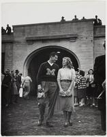 Image of a man and a woman outside a stone or brick archway. The woman wears a light-colored sweater and darker skirt and the man wears a sweater with a block &quot;M&quot; on it.