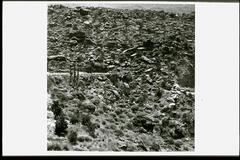 This is a black and white photograph of a rocky hillside filled with boulders and brush vegetation. In the distance there is more rocky terrain. In the middle area there is paved terrace and to the right a section of the hill has been cut away.