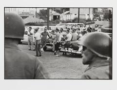 A black and white photograph of two helmeted figures in the foreground, and a crowd of people gathered around cars and looking to the viewer&#39;s left. Buildings and gravel piles appear in the background.