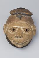 A mask with an oval shaped head and protruding features. The mouth features articulated teeth. A blue bird sits atop the head of the mask.&nbsp;