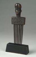 This wooden comb, or <em>cisakulo,</em> is composed of six long teeth and an anthropomorphic support. Its handle includes a rectangular section with multiple bands of diagonal, incised lines. This section along with the teeth of the comb visually form an abstracted body for the delicately carved head, which sits atop the handle. These lined motifs as well as the fine facial features are similar to those found in the figural carvings of the neighboring Chokwe; the striated turbanesque coiffure, however, is distinctly characteristic of the Lwena style.