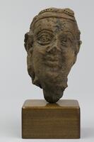 A detailed head featuring decorative dot inlays around a headpiece. The face is smiling and eyebrows are joined. The verso features vertical dashes for hair and the headpiece juts outward.&nbsp;