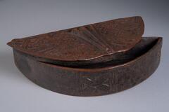Crescent shaped box with lid. Box contains various geometric patterns. The center of the lid is designed with three deeply carved lines. Covering the remainder of the lid, on both sides of the three lines, are diamond patterns formed by interlocking lines.