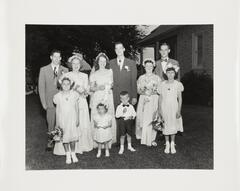 A black and white image of three men, one woman, five girls, and one boy standing in front of a house. The woman wears a veil and she and four girls hold bouquets. The men wear suits and boutonniers. The small girl and boy stand in front of the rest of the party. 