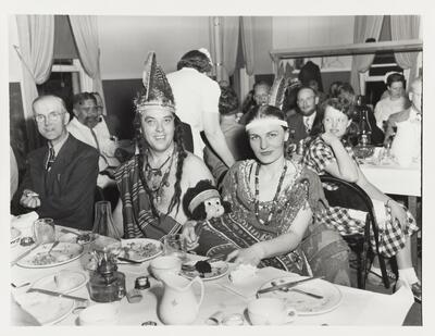A black and white image of a man and woman dressed as Native Americans at a dinner. The woman is holding a doll wrapped in a blanket. 